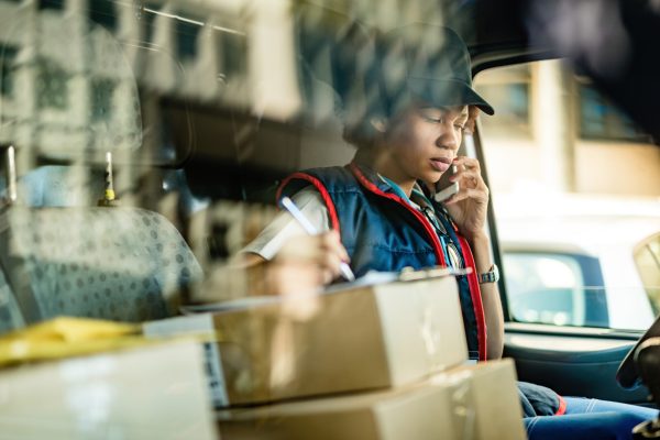 Black female courier communicating on mobile phone and taking notes while sitting in delivery van. The view is through window.